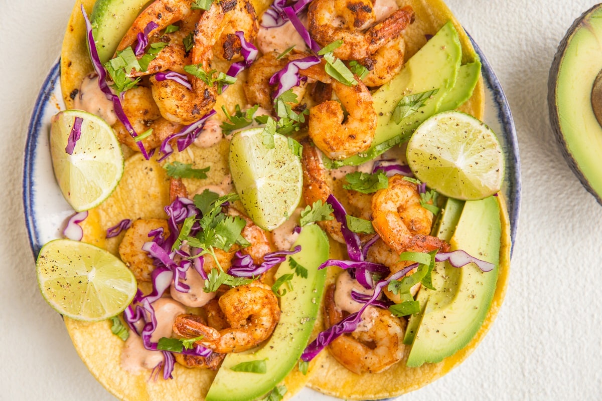 horitzonal image of a plate of shrimp tacos with slices of lime and chipotle sauce.