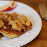 A white plate with pancakes topped with chunky cooked apples sits on a wooden table. A fork and a few slices of apple are next to the plate.