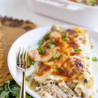 Plate with two chicken enchiladas on it with a golden fork and fresh cilantro to the side and the casserole dish with more enchiladas in the background.