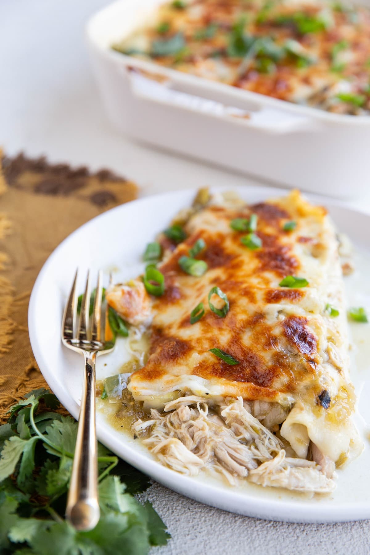 Plate with two chicken enchiladas on it with a golden fork and fresh cilantro to the side and the casserole dish with more enchiladas in the background.