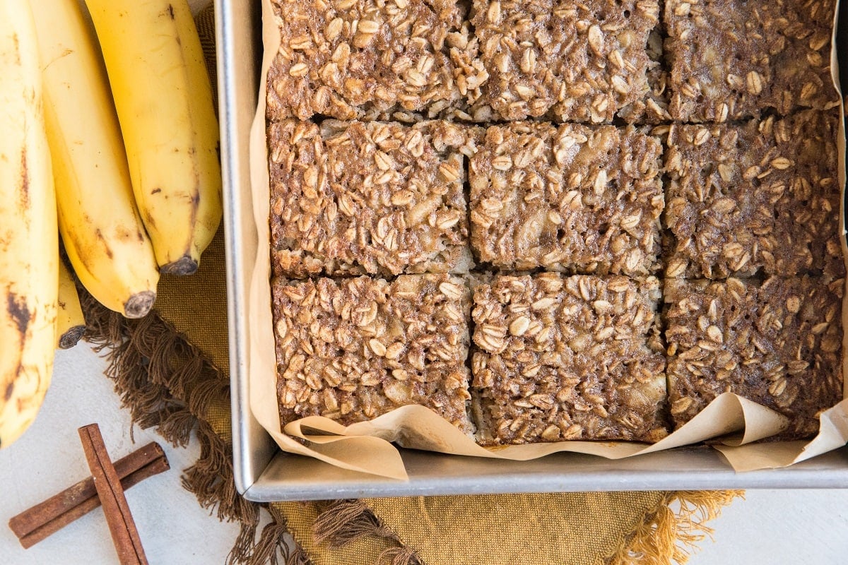 Horizontal photo of banana baked oatmeal in a baking pan sliced into squares with a bunch of bananas off to the side