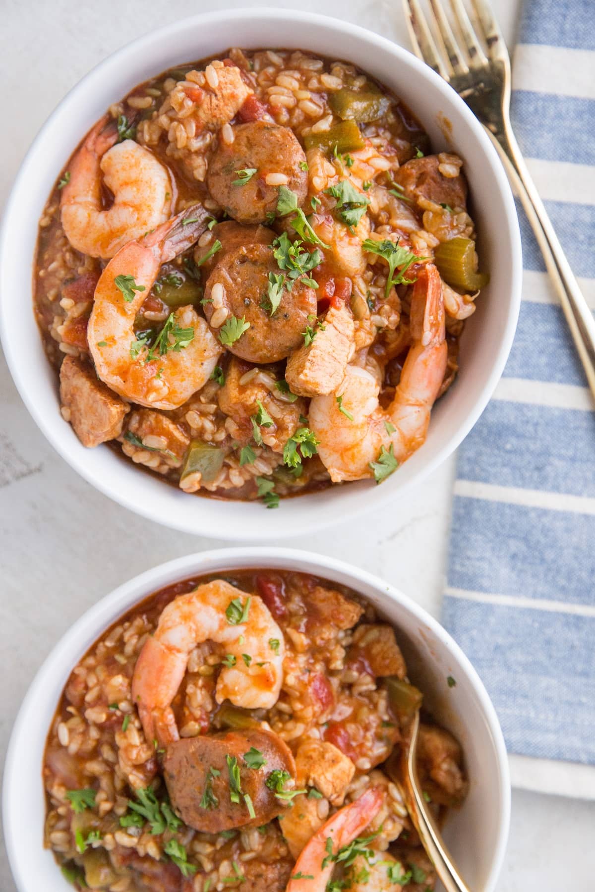 Top down photo of two white bowls of jambalaya with a blue striped napkin and a gold spoon in one of the bowls