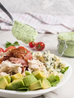 Cobb Salad with Homemade Green Goddess Dressing in a white bowl with green goddess dressing in the background on a marble surface.