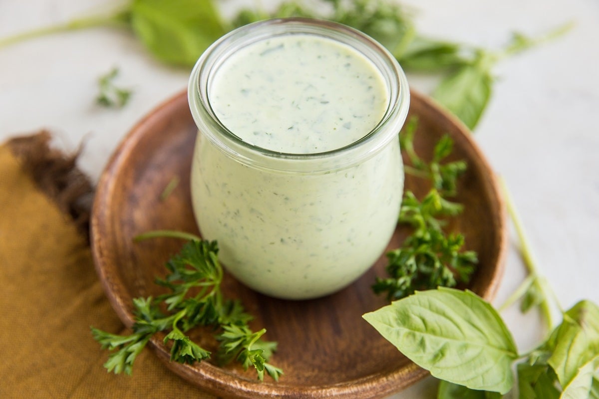 Mayo-Free Green Goddess Dressing sitting on a wooden plate in a jar with fresh herbs to the side and a golden napkin