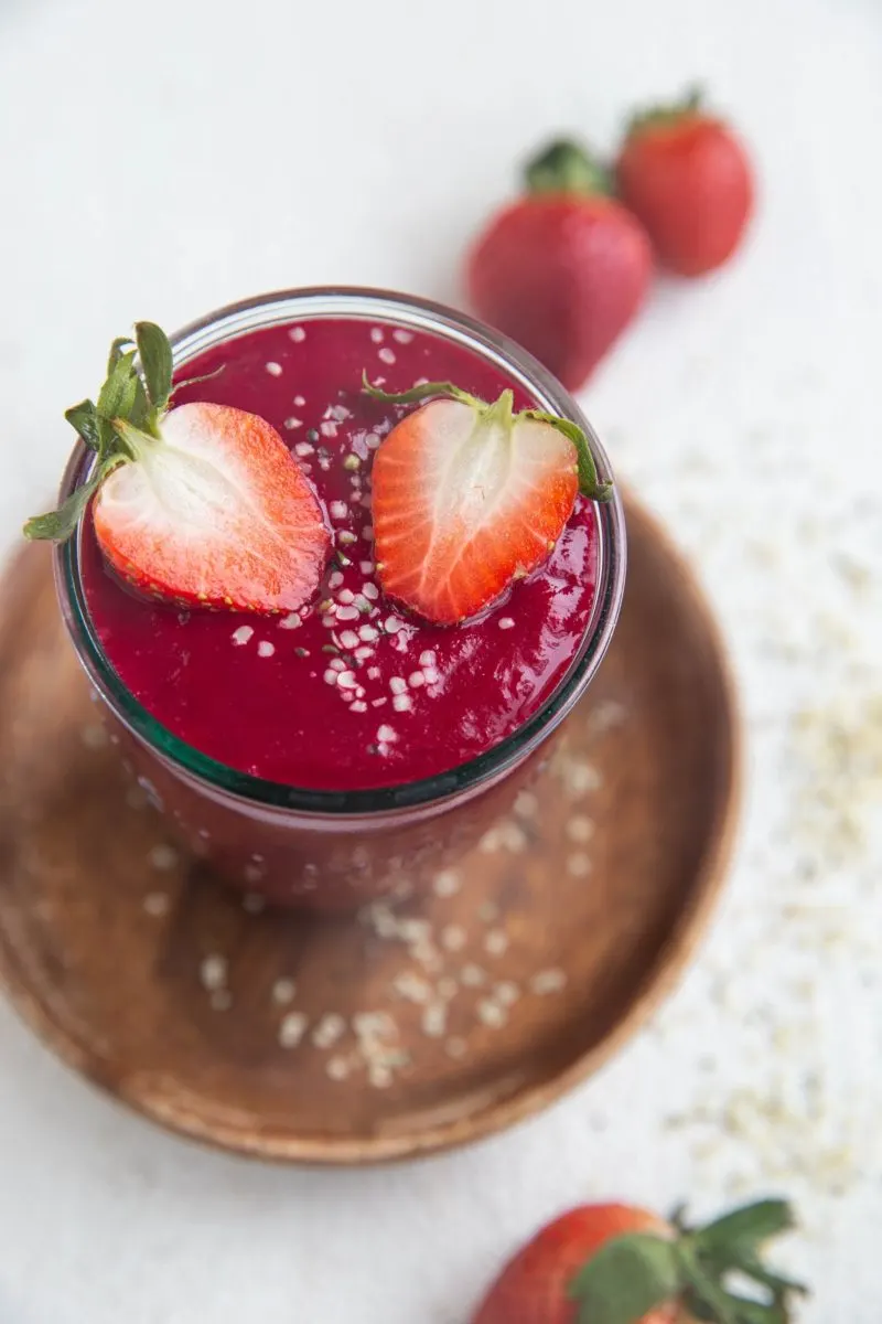 An anti-inflammatory beet smoothie that is deep red inside a glass with hemp seeds on top and sliced strawberries, on a white background.