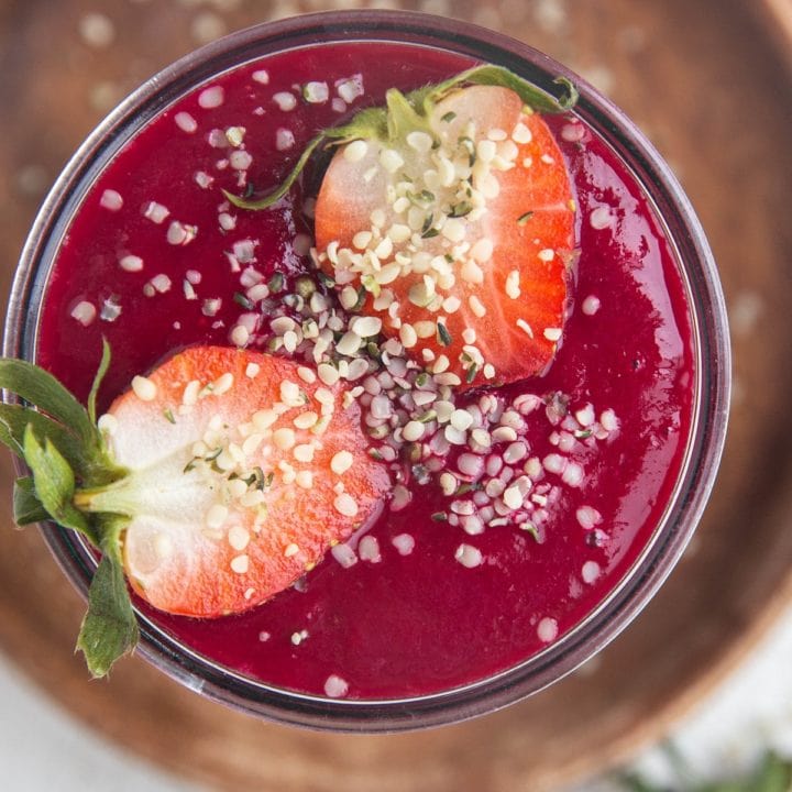 Close-up top down image of a red smoothie in a glass sitting on a wooden plate on a white background. Sprinkled with hemp seeds.