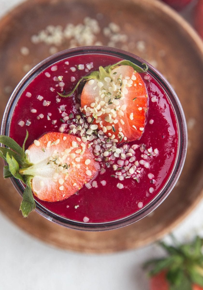 Close-up top down image of a red smoothie in a glass sitting on a wooden plate on a white background. Sprinkled with hemp seeds.