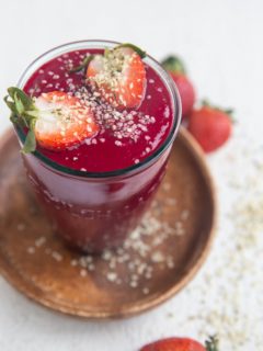 Glass of anti-inflammatory beet smoothie with a beautiful red color. The smoothie is sprinkled with hemp seeds and has a sliced strawberry on top.