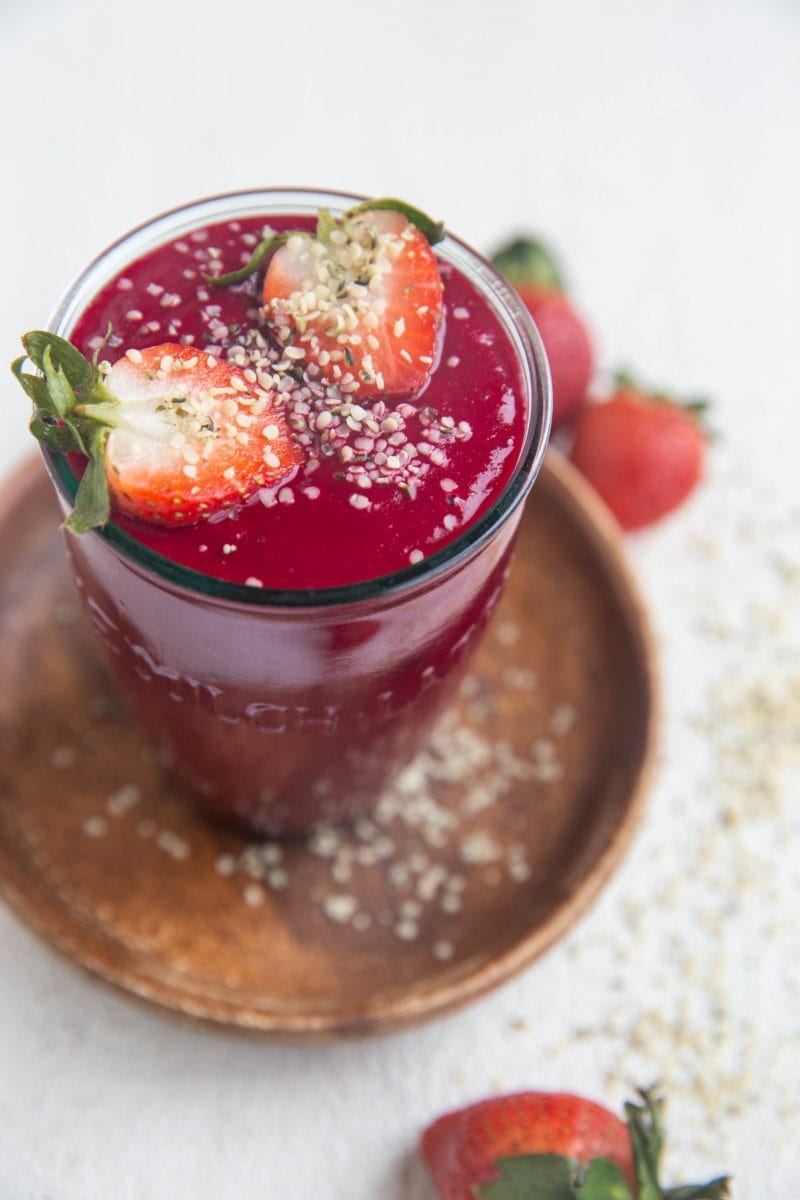 Glass of anti-inflammatory beet smoothie with a beautiful red color. The smoothie is sprinkled with hemp seeds and has a sliced strawberry on top.