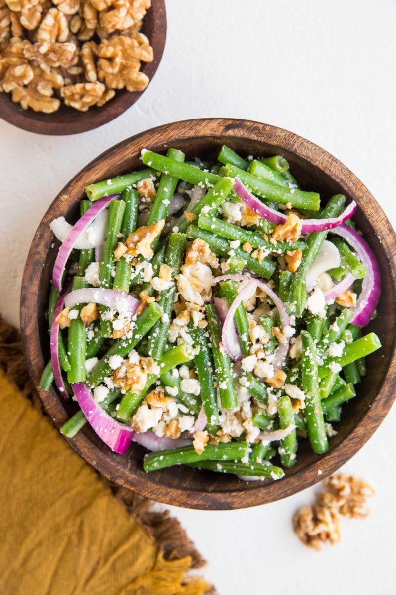top down photo of green bean salad in a wooden bowl with a golden napkin to the side and walnuts in a bowl.