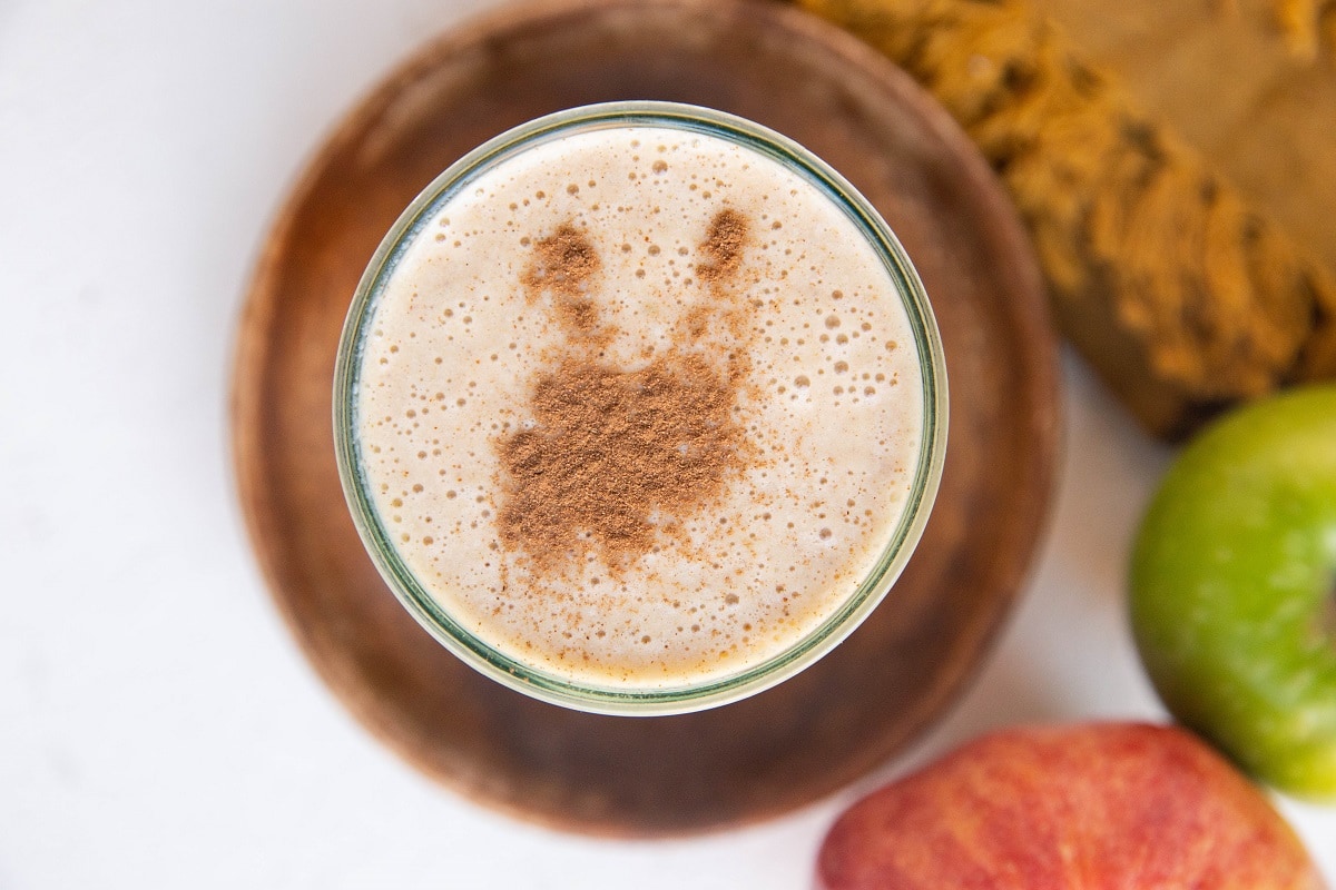 Horizonal top down photo of an apple smoothie in a glass sitting on a wooden round plate with fresh apples next to it.