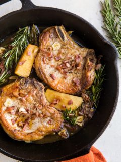 Roasted Pork Chops with cider-spiced apples, bacon, roasted garlic, and rosemary in a cast iron skillet on top of a white backdrop with a red napkin