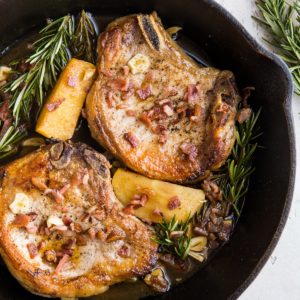 Roasted Pork Chops with cider-spiced apples, bacon, roasted garlic, and rosemary in a cast iron skillet on top of a white backdrop with a red napkin