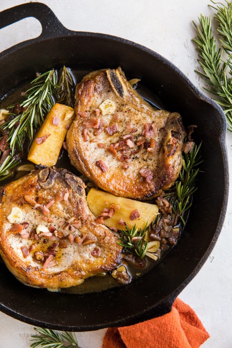 Roasted Pork Chops with cider-spiced apples, bacon, roasted garlic, and rosemary in a cast iron skillet on top of a white backdrop with a red napkin