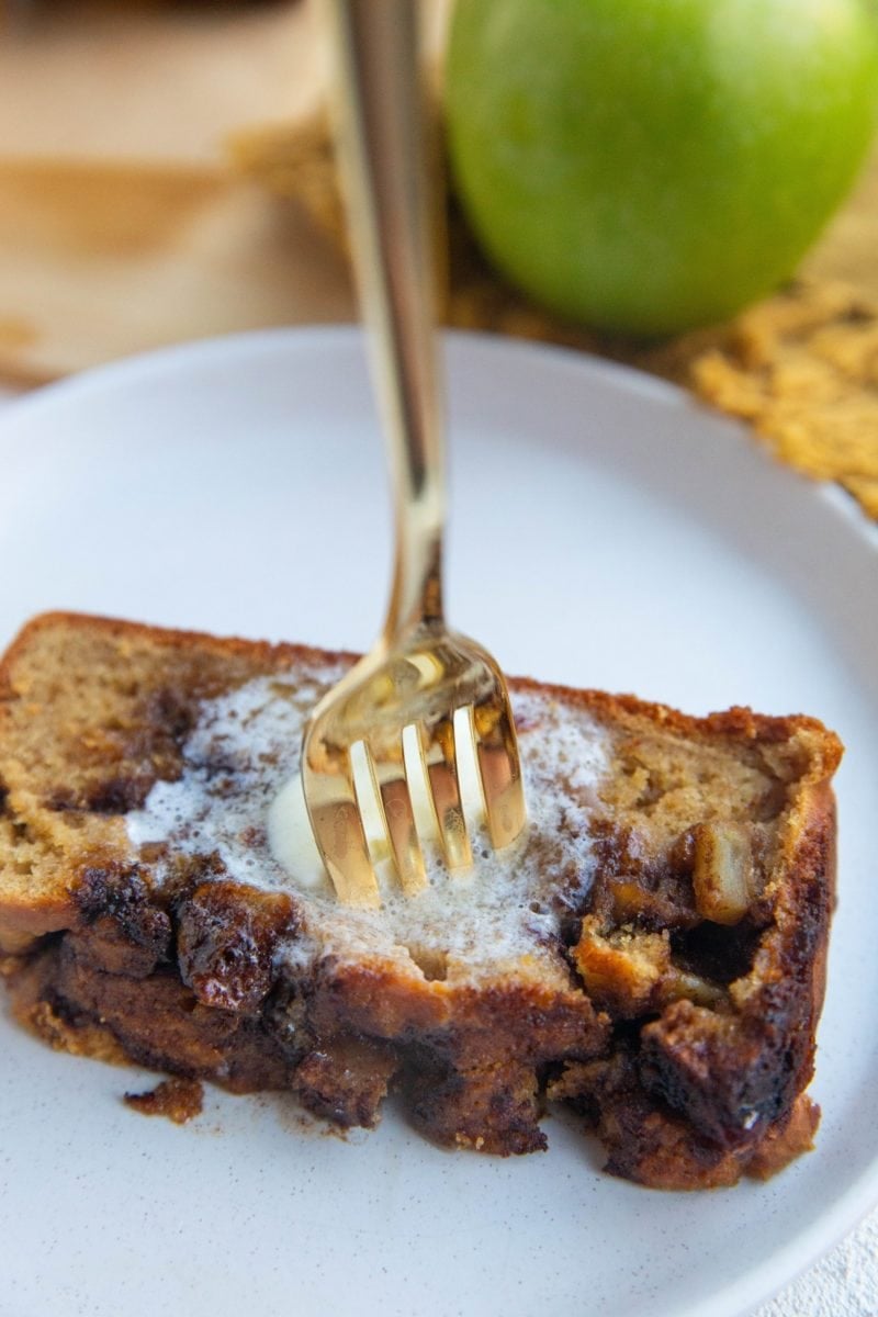 Slice of apple bread on a plate with melted butter on top and a fork taking a bite.