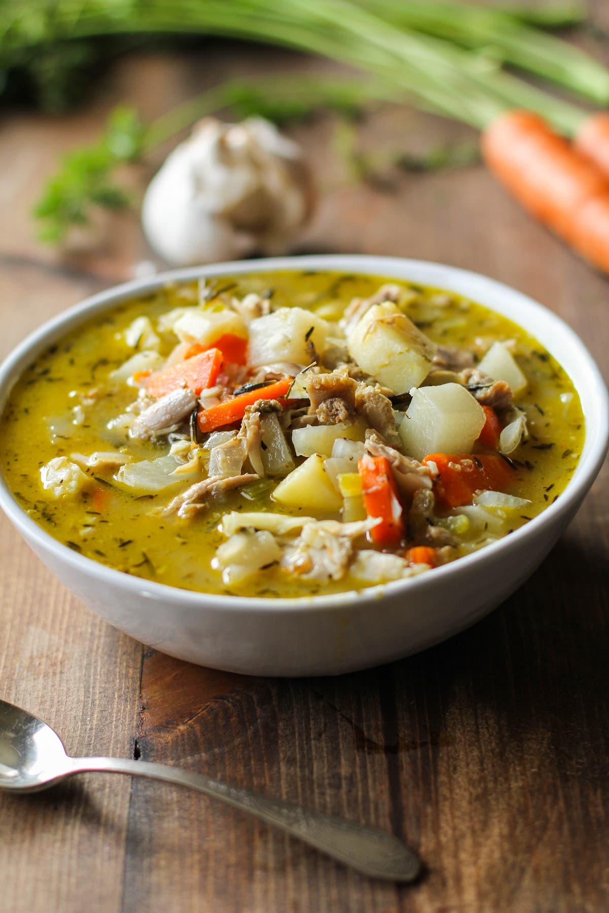 Bowl of turkey soup with a spoon to the side and vegetables in the background on a wood backdrop.