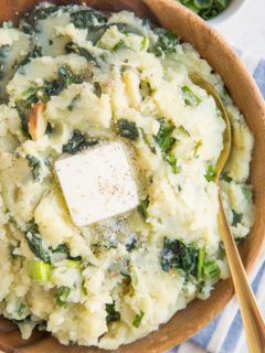 Close up image of colcannon in a wooden bowl with a blue striped napkin, a gold spoon and melted butter on top of the potatoes