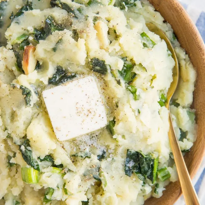 Close up image of colcannon in a wooden bowl with a blue striped napkin, a gold spoon and melted butter on top of the potatoes