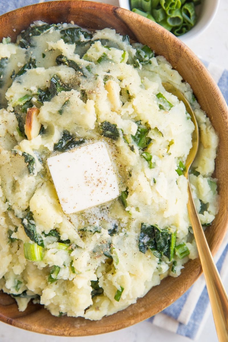 Close up image of colcannon in a wooden bowl with a blue striped napkin, a gold spoon and melted butter on top of the potatoes