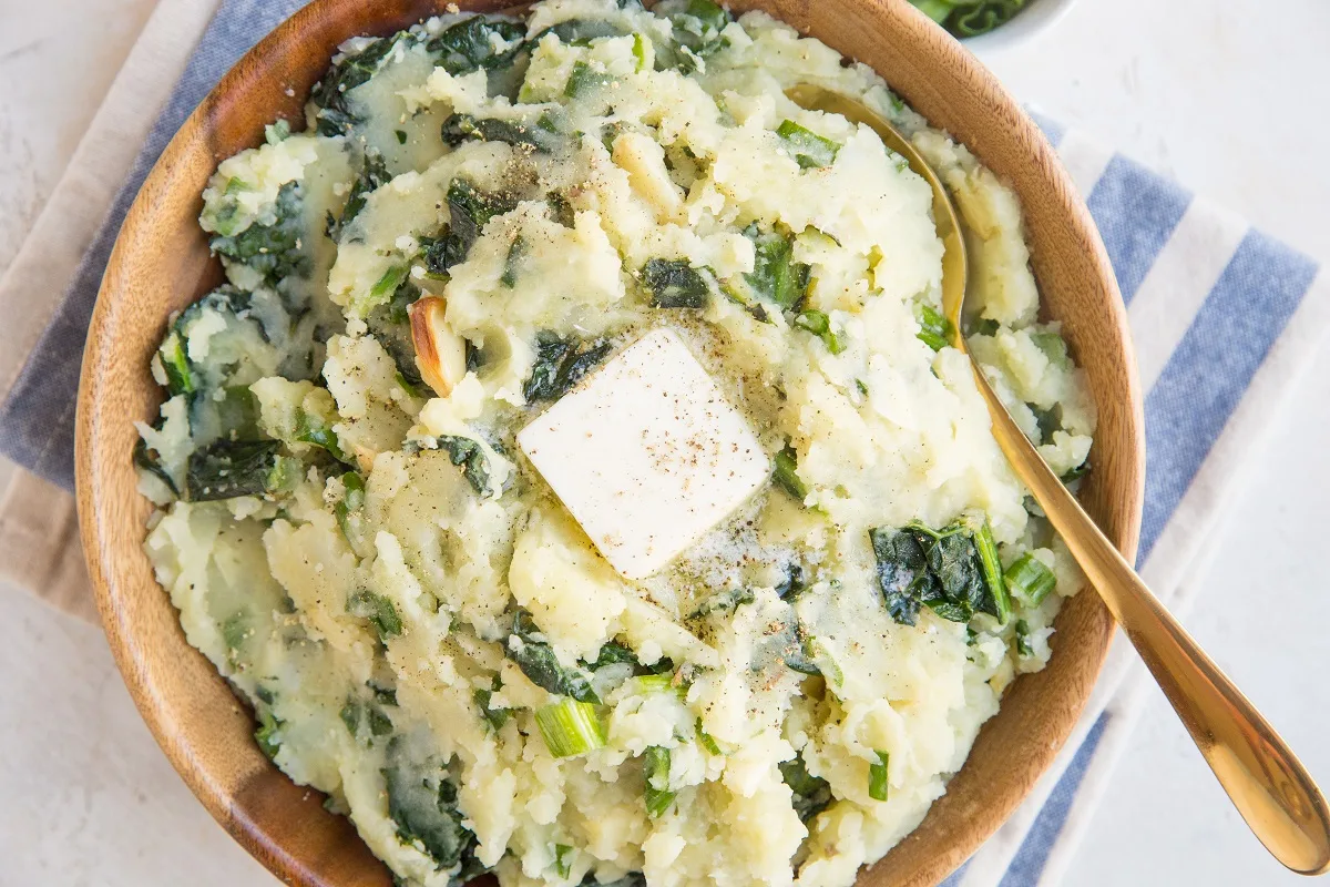 Colcannon in a large wooden bowl with butter and a gold spoon. Horizontal photo with blue napkin under bowl of mashed potatoes