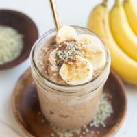 Mason jar of overnight oatmeal with sliced banana, almond butter, and hemp seeds on top with a gold spoon.