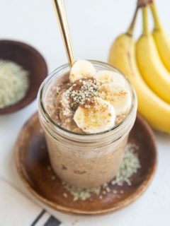 Mason jar of overnight oatmeal with sliced banana, almond butter, and hemp seeds on top with a gold spoon.