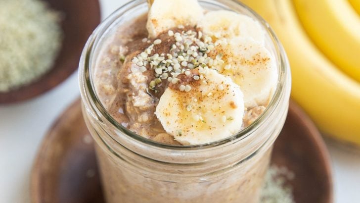 Mason jar of overnight oatmeal with sliced banana, almond butter, and hemp seeds on top with a gold spoon.