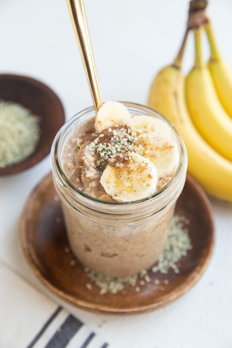 Mason jar of overnight oatmeal with sliced banana, almond butter, and hemp seeds on top with a gold spoon.