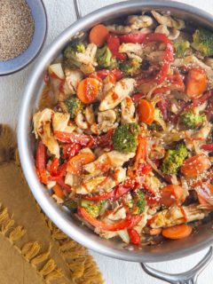 Top down photo of a stainless steel skillet full of chicken stir fry with vegetables. A golden napkin and a bowl of sesame seeds to the side.