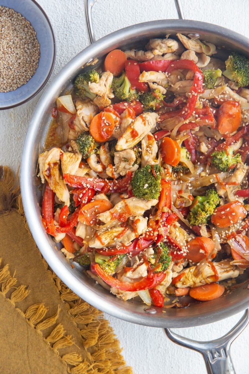 Top down photo of a stainless steel skillet full of chicken stir fry with vegetables. A golden napkin and a bowl of sesame seeds to the side.