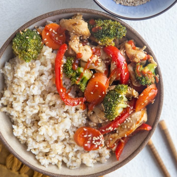Top down photo of a ceramic bowl full of steamed brown rice and healthy chicken stir fry with veggies. Chopsticks and a gold napkin to the side.