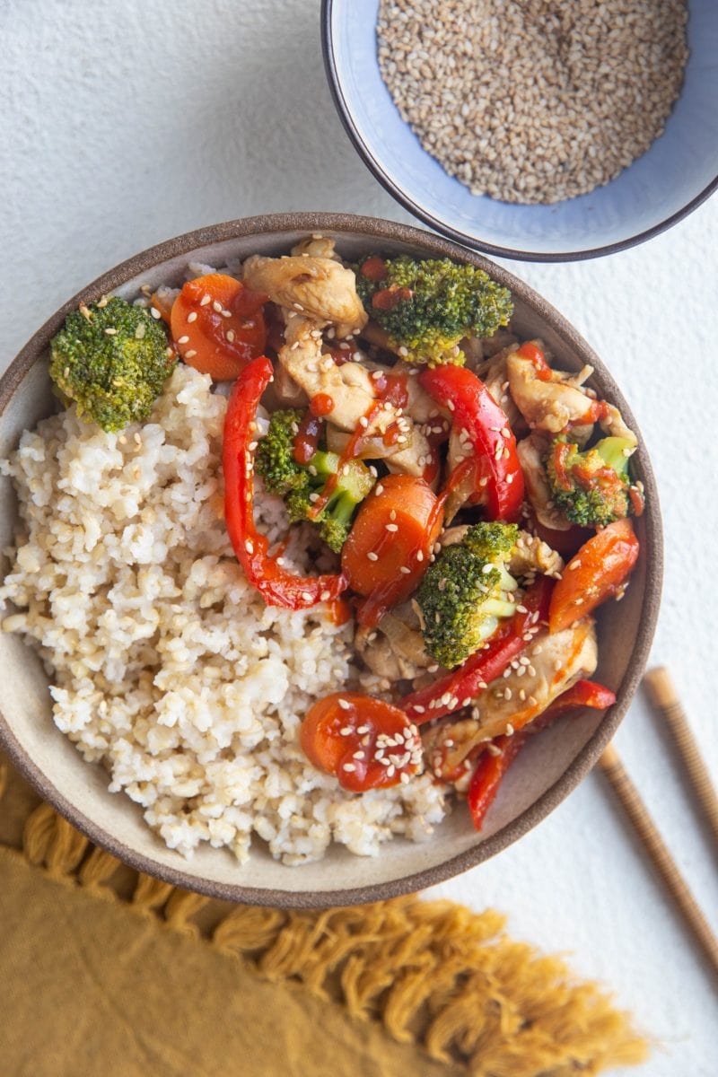 Top down photo of a ceramic bowl full of steamed brown rice and healthy chicken stir fry with veggies. Chopsticks and a gold napkin to the side.