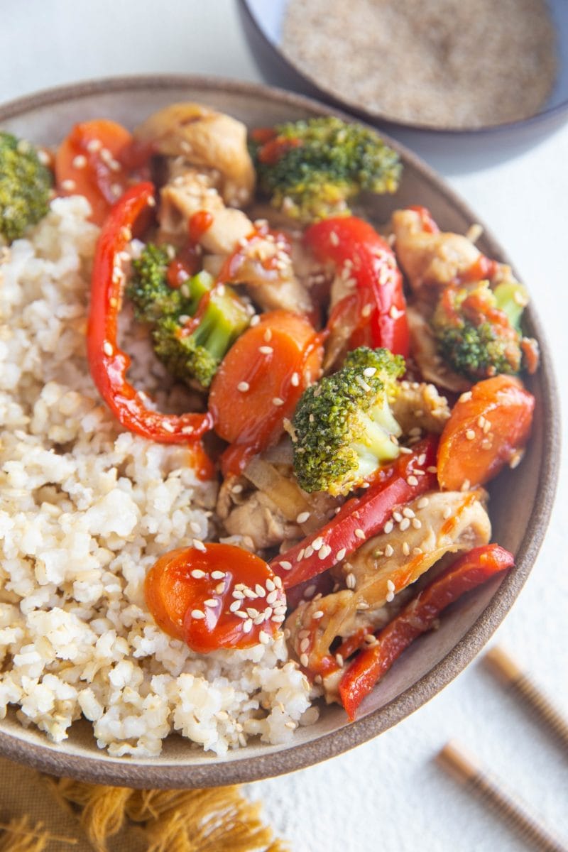 Angled photograph of a big bowl of brown rice and chicken stir fry, ready to eat. Chopsticks and a napkin to the side.