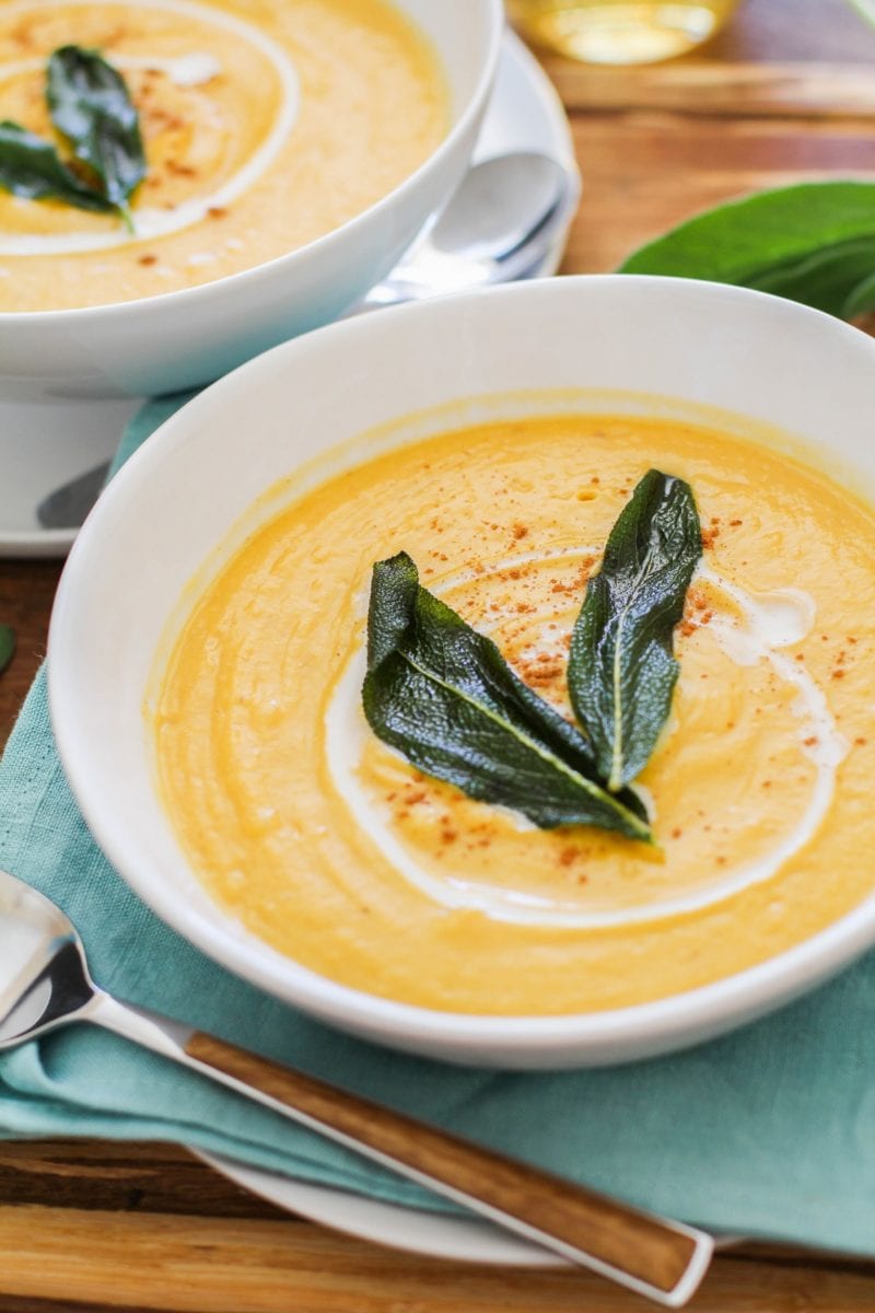 Two white bowls of butternut squash soup on a wooden cutting board with a blue napkin and wood inlay spoons and wine.