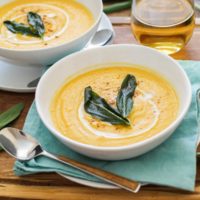 Two bowls of butternut squash soup with crispy sage on top, sitting on top of a wooden cutting board on a wood rustic backdrop. Blue napkin, wood inlay spoons, and white wine.