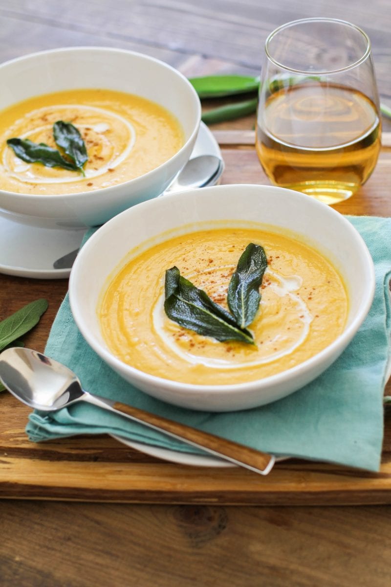 Two bowls of butternut squash soup with crispy sage on top, sitting on top of a wooden cutting board on a wood rustic backdrop. Blue napkin, wood inlay spoons, and white wine.
