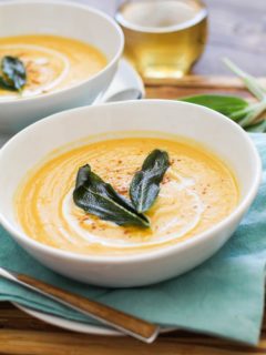 Two white bowls with butternut squash soup on a wooden cutting board with wood inlay spoons and white wine in the background.