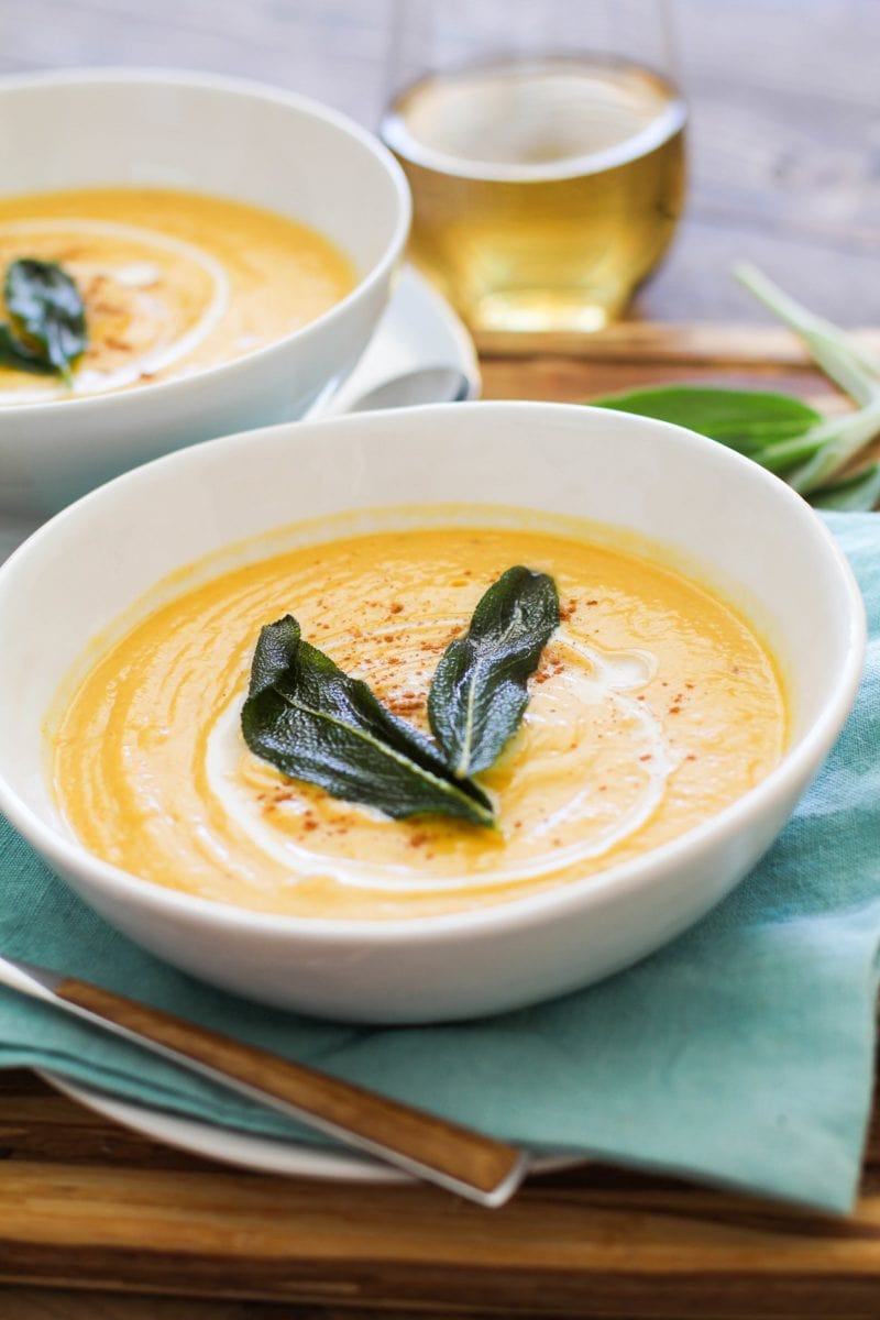 Two white bowls with butternut squash soup on a wooden cutting board with wood inlay spoons and white wine in the background.