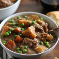 two white bowls of hearty beef stew on a rustic wood background with bread and wine to the side.