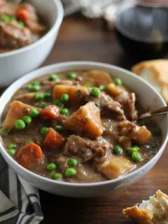 two white bowls of hearty beef stew on a rustic wood background with bread and wine to the side.