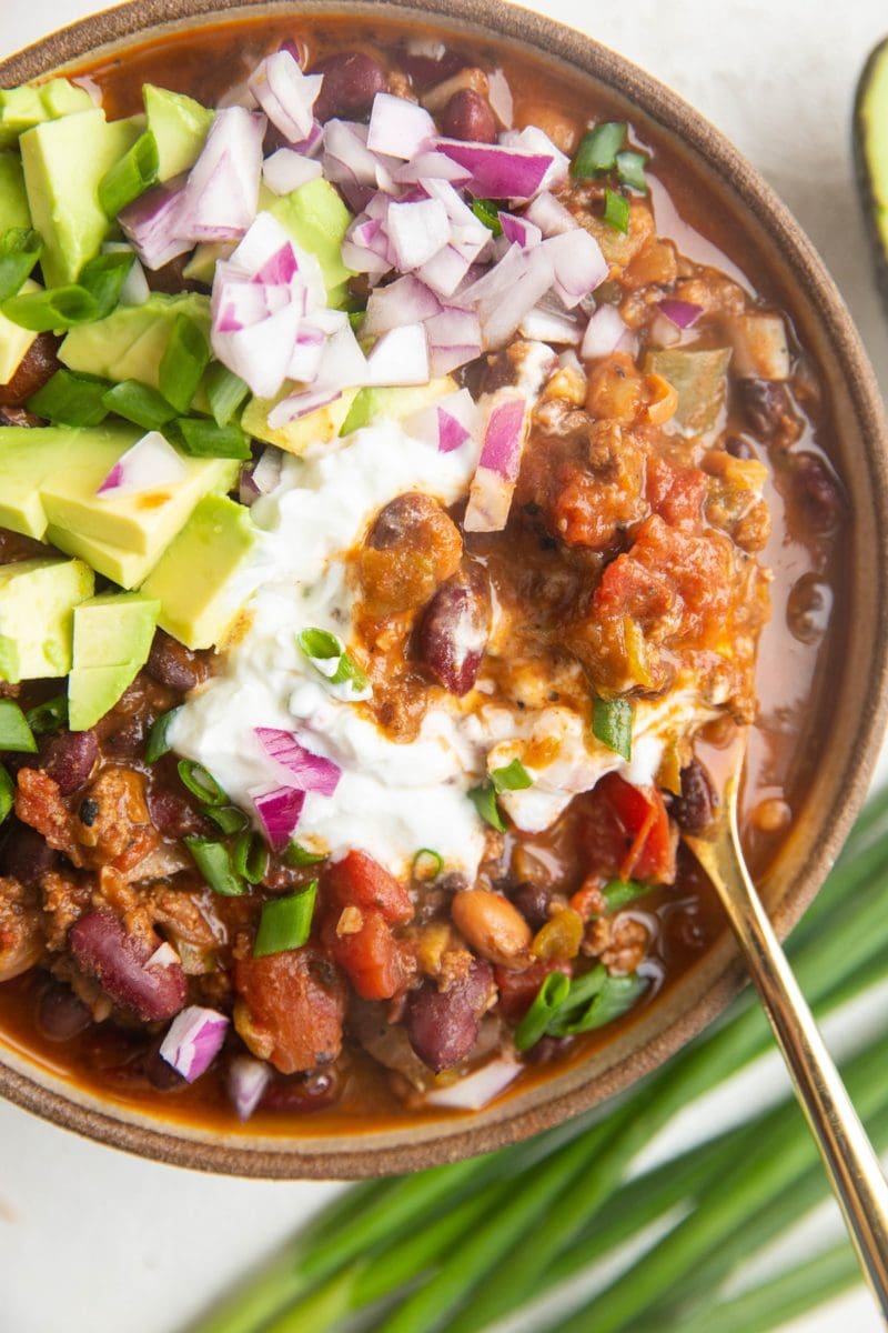 Close up top down photo of a big bowl of turkey chili with sour cream, diced avocado, red onion and green onion with a gold spoon, ready to eat.