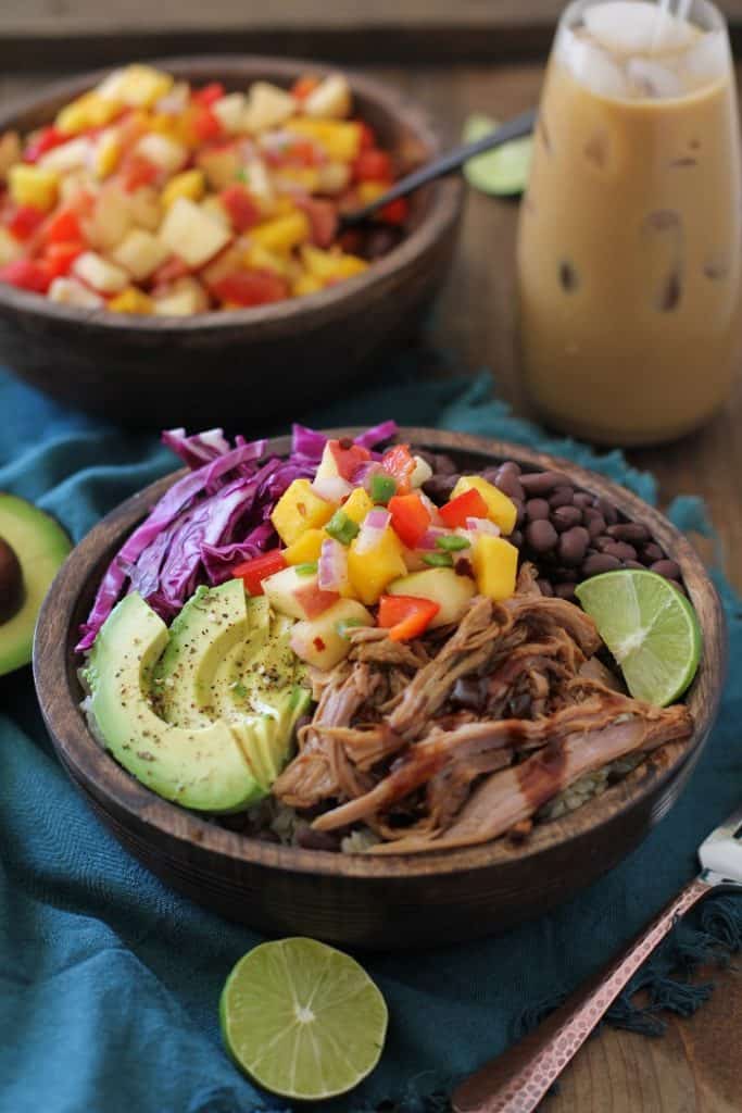 A colorful bowl with crock pot Hawaiian pulled pork, sliced avocado, black beans, purple cabbage, lime, and pineapple salsa over quinoa. An iced coffee and another bowl of salsa are in the background.