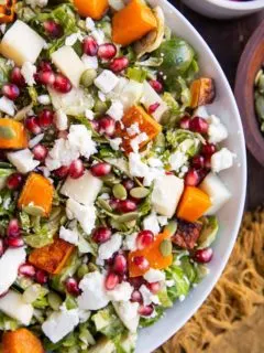 Close up top down photo of white bowl of Shaved Brussel Sprout Salad with Roasted Butternut Squash, Pomegranate Seeds, Pumpkin Seeds, Feta, and Citrusy Maple Cinnamon Dressing