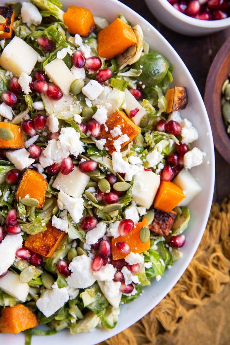Close up top down photo of white bowl of Shaved Brussel Sprout Salad with Roasted Butternut Squash, Pomegranate Seeds, Pumpkin Seeds, Feta, and Citrusy Maple Cinnamon Dressing