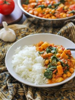 White bowl with chana saag and white rice inside. An ornate decorative napkin underneath the bowl and a skillet full of chickpea curry in the background.