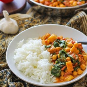 White bowl with chana saag and white rice inside. An ornate decorative napkin underneath the bowl and a skillet full of chickpea curry in the background.