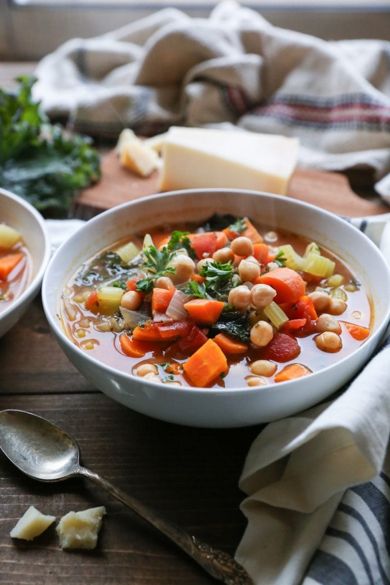 Big white bowl of minestrone soup with hunks of parmesan, a spoon and napkin to the side.