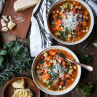 Rustic Minestrone Soup with Rice and Kale in two white bowls on a wooden backdrop with a plate of cornbread to the side.