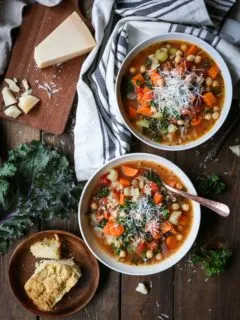 Rustic Minestrone Soup with Rice and Kale in two white bowls on a wooden backdrop with a plate of cornbread to the side.