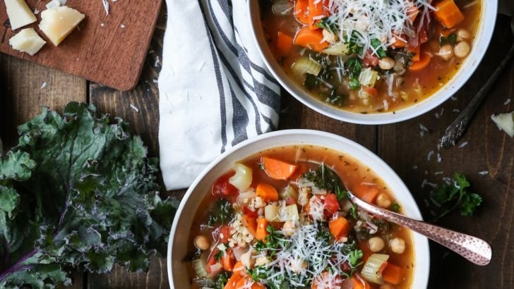 Rustic Minestrone Soup with Rice and Kale in two white bowls on a wooden backdrop with a plate of cornbread to the side.